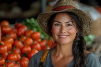 Smiling Hispanic female farmer in straw hat at farmers market stand with fresh red tomatoes. Natural lighting highlights authentic farm-to-table atmosphere.