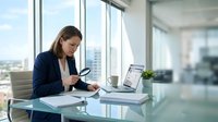 Professional businesswoman using smartphone at modern office desk with laptop and documents in bright corporate workspace with city view.