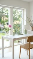 Bright minimalist dining room with white table, wooden chair, fresh flowers in vase, and large windows overlooking garden for modern home interior design.