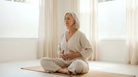 Mature Caucasian woman practicing meditation in lotus position on yoga mat in bright sunlit room wearing comfortable beige clothing for wellness and mindfulness.