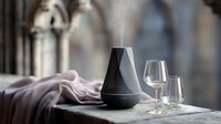 Modern black aroma diffuser with empty wine glasses on marble surface against blurred gothic window background, creating luxurious spa atmosphere.
