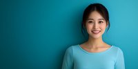 Young Asian woman in light blue top smiling warmly against turquoise wall background, natural makeup and casual style for lifestyle, beauty, and wellness concepts.