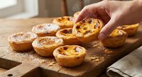 Hand picking golden Portuguese egg tart from wooden board with freshly baked pasteis de nata custard pastries dusted with cinnamon in warm kitchen setting.