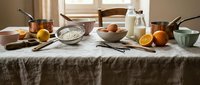 Rustic kitchen table with baking ingredients flour eggs milk oranges and copper cookware on linen tablecloth for homemade cooking and recipe preparation.