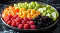 Fresh colorful fruit platter with raspberries, blackberries, mango cubes, kiwi slices and green grapes arranged in rainbow pattern on dark plate, healthy food.