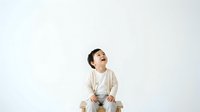 Young toddler with dark hair, appearing to be of Asian ethnicity, wearing casual cream and white clothing, sitting and looking upward with curious expression against white background.