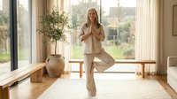 Caucasian woman in beige wellness attire practicing yoga tree pose in bright modern living room with large windows, wooden floors, and natural light for mindfulness and fitness content.