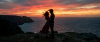 Silhouetted couple embracing on rocky coastal cliff at golden sunset with ocean and dramatic sky backdrop for romance, love, and travel concepts.