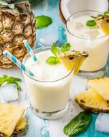 Refreshing pineapple coconut smoothie with mint garnish and striped straws on rustic turquoise background, surrounded by fresh fruit slices and ice cubes.