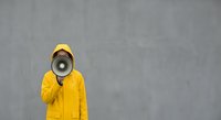 Person in bright yellow raincoat and cap holding megaphone against concrete wall background for communication and announcement concepts.