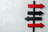 Red and black directional arrow signs on wooden post against white textured wall showing multiple path options for decision making and navigation concepts.