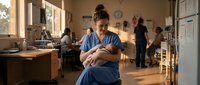 African American female nurse in blue scrubs holding newborn baby in hospital maternity ward with medical staff working in background during golden hour lighting.