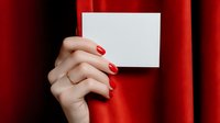 Female hand with red manicure holding blank white card against luxurious red velvet curtain background.