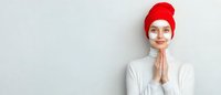 Young woman in red beanie and white sweater with hands clasped in prayer or gratitude pose against light background, expressing peaceful mindful moment.