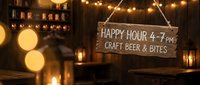 Rustic wooden happy hour sign advertising craft beer and bites from 4-7pm hanging in warm cozy bar atmosphere with string lights and bokeh background.
