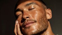 Young Hispanic man with closed eyes and glowing skin touching face against dark background, showcasing skincare routine or wellness moment.