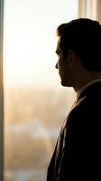Silhouette of contemplative businessman in suit looking out office window during golden hour light for corporate and professional concepts.