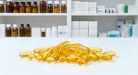 Golden omega-3 fish oil capsules scattered on white surface with blurred pharmacy shelves background showing medicine bottles and pharmaceutical products.