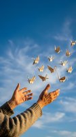 Hands releasing golden origami paper cranes into blue sky with clouds symbolizing freedom hope peace and spiritual transformation.