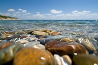 Smooth pebbles underwater at beach shoreline with crystal clear water and blue sky, split level view showing both above and below water surface perspective.