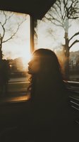 Woman with long dark hair in profile, silhouetted against golden sunset light streaming through window with bare tree branches visible. Contemplative mood suitable for wellness, mental health, lifestyle and editorial projects.