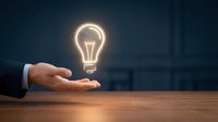 Male hand in business suit holding glowing neon lightbulb on wooden table against dark background, representing innovation, creativity, ideas and business solutions.