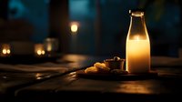 Glass bottle of fresh milk with cookies on wooden table in dark moody setting with warm ambient lighting creates cozy atmosphere for food photography and advertising.