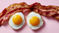 Crispy fried bacon strips and sunny side up eggs with bright yellow yolks on pink background. Classic breakfast composition for food photography and menu design.