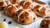 Freshly baked hot cross buns with raisins on parchment paper, golden brown with white icing crosses, traditional Easter pastry.