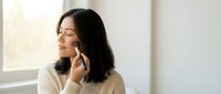 Young Asian woman in beige sweater talking on smartphone by window with natural light, peaceful expression for communication and lifestyle content.