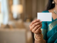 Woman in teal attire holding blank white card with gold bracelets, indoor setting with warm blurred background, suitable for business cards and promotional materials.