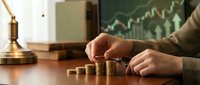 Professional businessman stacking golden coins on wooden desk with financial growth chart background for investment and wealth management concepts.