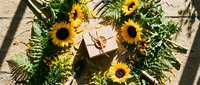 Sunflowers and ferns arranged around wrapped gift box on textured surface with natural shadows creating rustic botanical flat lay composition for celebrations.
