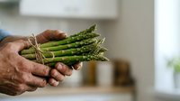 Fresh green asparagus bunch held in male hands with blurred kitchen background, suitable for cooking, recipe, and food preparation content.