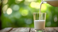 Fresh milk being poured into glass on wooden table with green bokeh background for healthy breakfast and dairy nutrition concepts.
