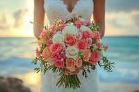 Bride holding romantic coral and white rose wedding bouquet against ocean sunset backdrop, featuring mixed roses and delicate greenery for beach ceremony.