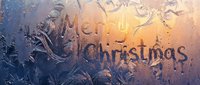 Merry Christmas text written on frosted winter window glass with ice patterns and warm golden light glowing through for holiday greeting cards.