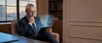 Senior male professional with gray hair and glasses reviewing digital holographic blueprint display on tablet in modern office environment, representing technology innovation and business planning.