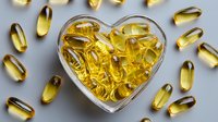 Heart-shaped glass bowl filled with golden omega-3 fish oil capsules, surrounded by additional supplements on gray background. Health concept.