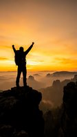 Silhouette of victorious hiker with raised arms standing on mountain peak at golden sunrise overlooking misty valley landscape for motivation and achievement concepts.