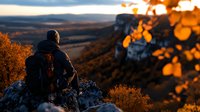 Person in warm clothing sitting on mountain peak at sunset, overlooking dramatic valley landscape with golden autumn foliage and rocky cliffs in warm evening light.