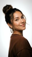 Young Hispanic woman with messy bun hairstyle wearing brown casual top and hoop earrings smiling at camera against white background for lifestyle content.