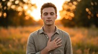 Young Caucasian man with light brown hair wearing casual linen shirt, hand on chest, standing in golden hour field at sunset with warm bokeh background.