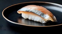 Fresh mackerel nigiri sushi piece on black ceramic plate with gold rim, showcasing traditional Japanese culinary artistry against dark background.