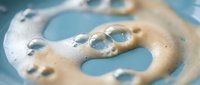 Creamy foam swirls with bubbles on a soft blue surface, capturing the texture of soap lather, skincare product, or beverage froth in a macro shot.