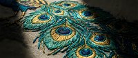 Intricate peacock feather embroidery with vibrant turquoise and gold threads on dark fabric showcasing detailed needlework and luxurious textile artistry.