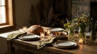 Rustic dining table set with ceramic plates, fresh bread, wildflowers in glass vase, and warm natural lighting creating cozy farmhouse atmosphere.
