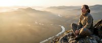 Middle-aged man with long hair and beard, Caucasian ethnicity, sitting on mountain peak at sunrise overlooking misty valley with winding river below. Adventure and contemplation theme for travel, lifestyle, and outdoor content.