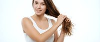 Young woman with long brown hair and fair complexion examining damaged hair ends against white background, representing hair care and beauty concerns.