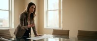 Professional businesswoman in suit standing at conference table using smartphone in modern office with natural lighting for corporate communications.
